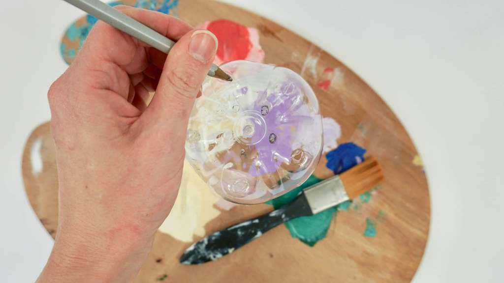 A close up of a hand holding a paintbrush over a water bottle on top of a paint palette.