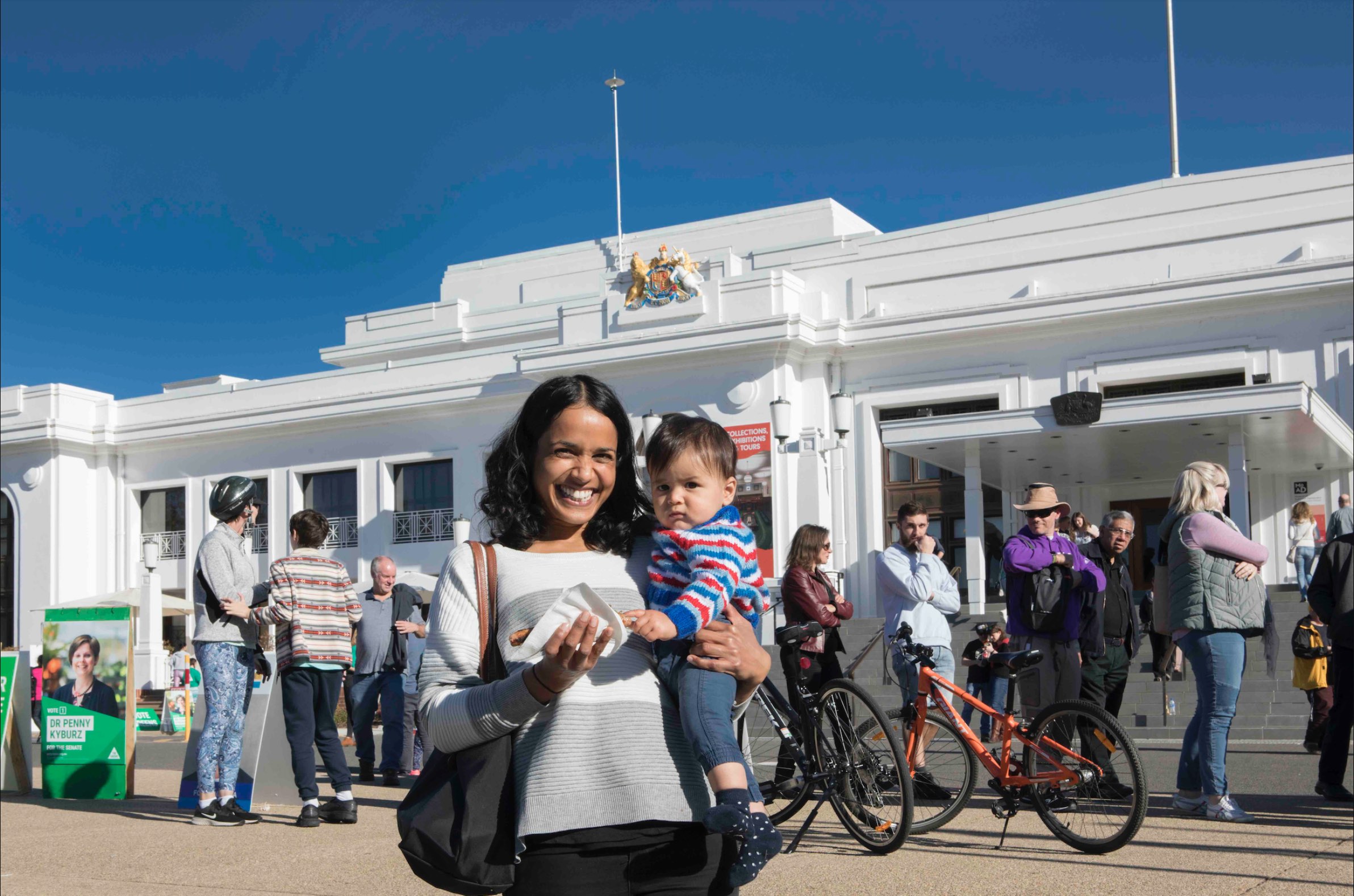 A mother holds her baby and smiles at the camera at the front of Old Parliament House. A row of people waiting to vote are behind her. She holds a sausage sandwich.