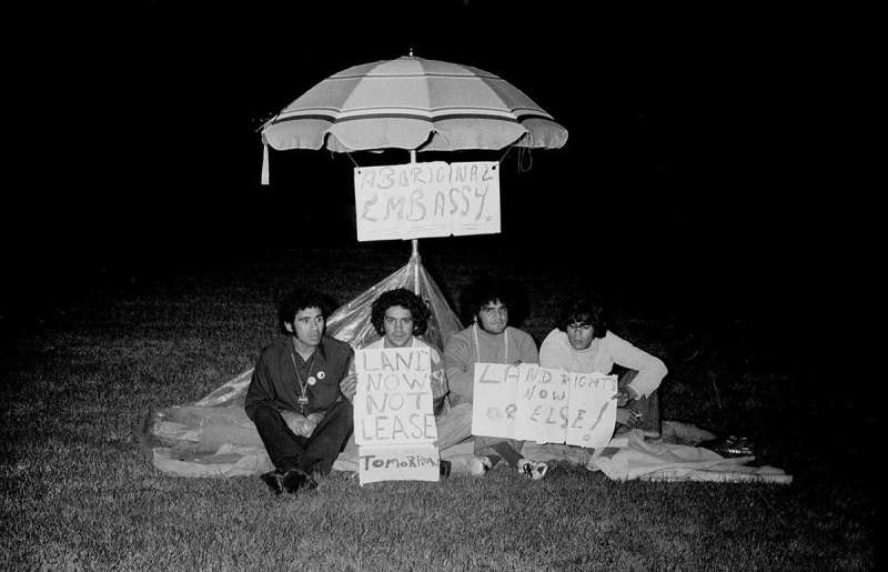 Four men sit on the ground under a beach umbrella with signs that read, 'Aboriginal Embassy', 'Land now not lease tomorrow' and 'Land rights now or else!'.