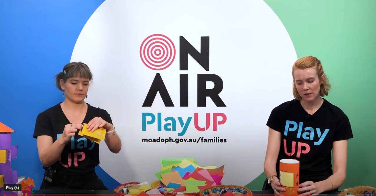 Two facilitators wearing black shirts that say PlayUP make something out of cardboard.