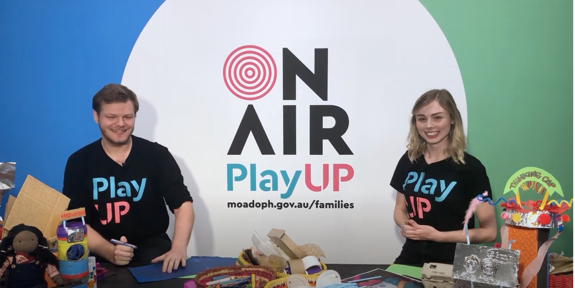 Two PlayUP facilitators stand behind a table covered in craft supplies.