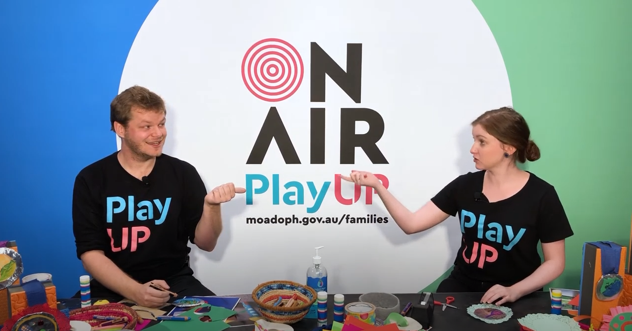 Two facilitators wearing black shirts that say PlayUP make something out of cardboard.