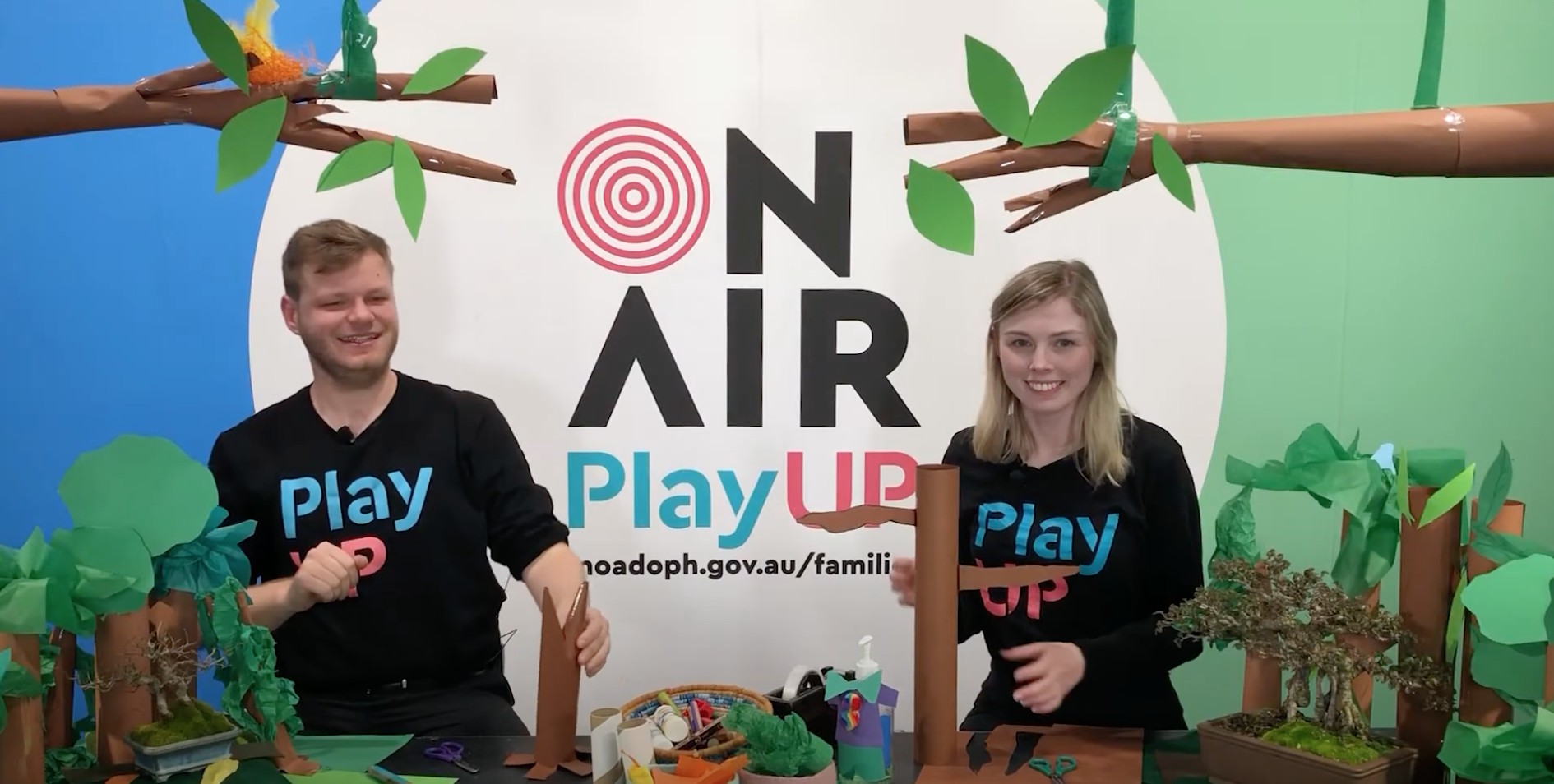 Two PlayUP facilitators are surrounded by paper trees and craft supplies. 