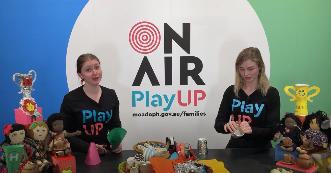 Two facilitators wearing black shirts that say PlayUP make something out of cardboard.