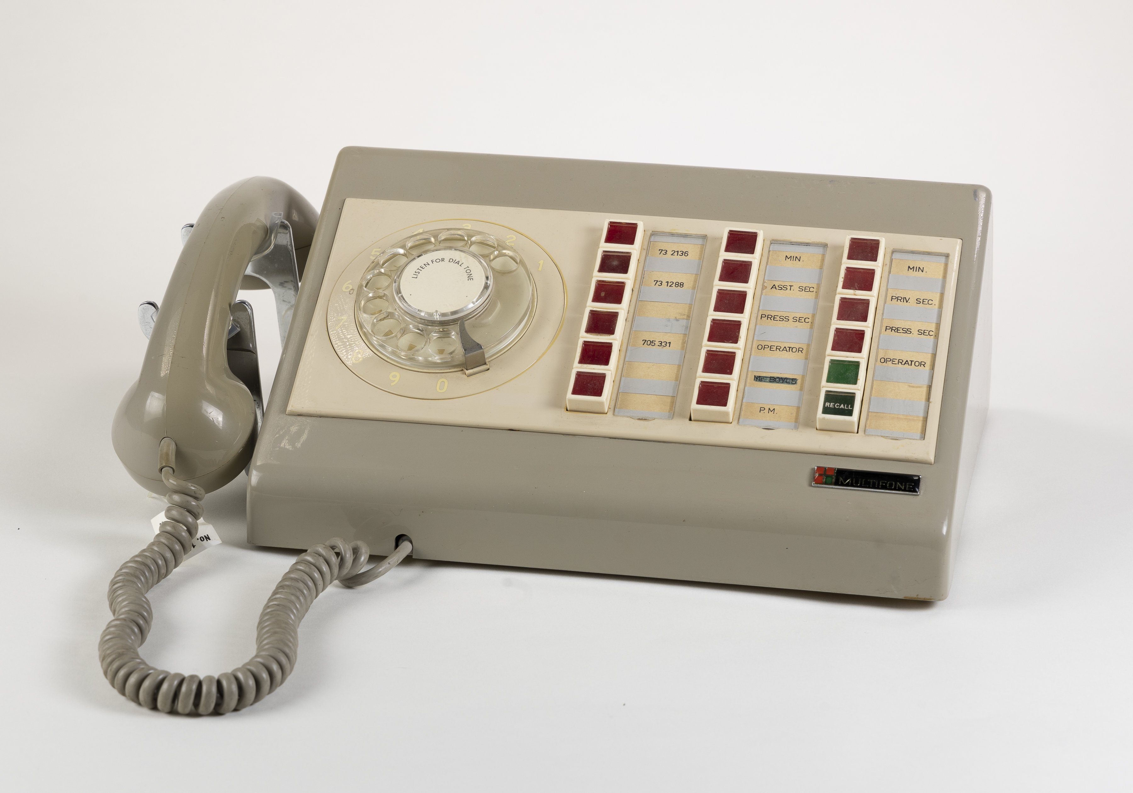 A light brown plastic phone with rows of red buttons and a manual hand dial. The receiver sits on the side. 