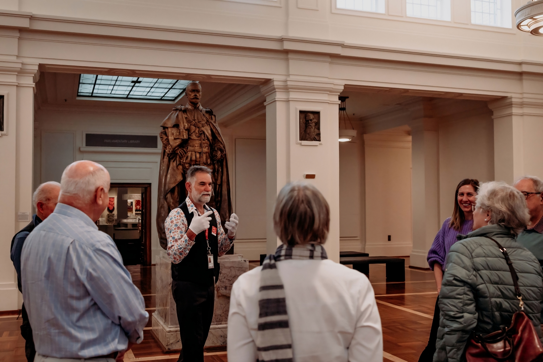 A tour group stand in a circle around a tour guide who wears white gloves and stands in front of a bronze statue. A tour group stand in a circle around a tour guide who wears white gloves and stands in front of a bronze statue.