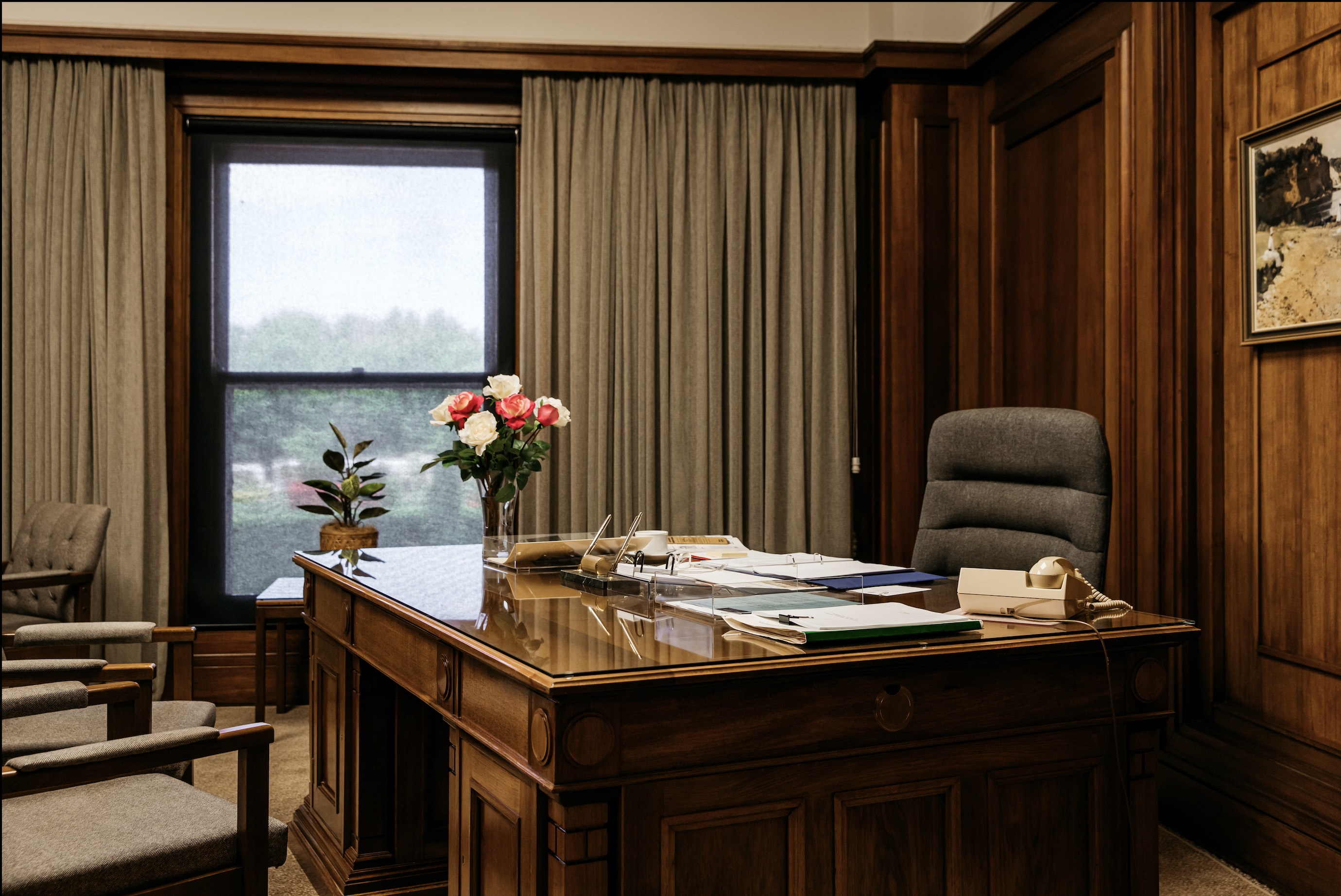 A large timber desk covered in folders and notes, with a vase with roses on it. 