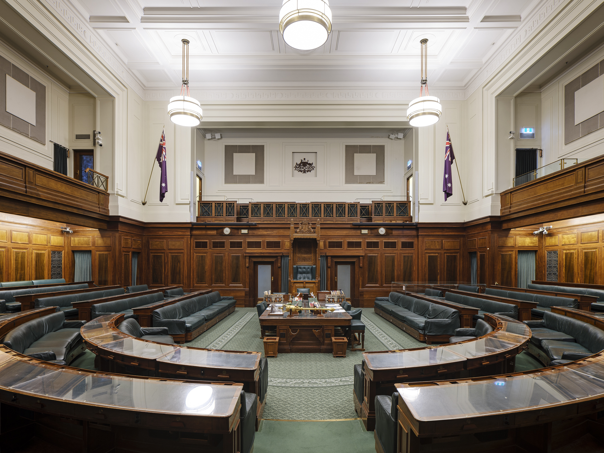 A green carpeted room with rows of wooden benches in a U shape facing a central table and the large, ornate Speaker's Chair. A green carpeted room with rows of wooden benches in a U shape facing a central table and the large, ornate Speaker's Chair.