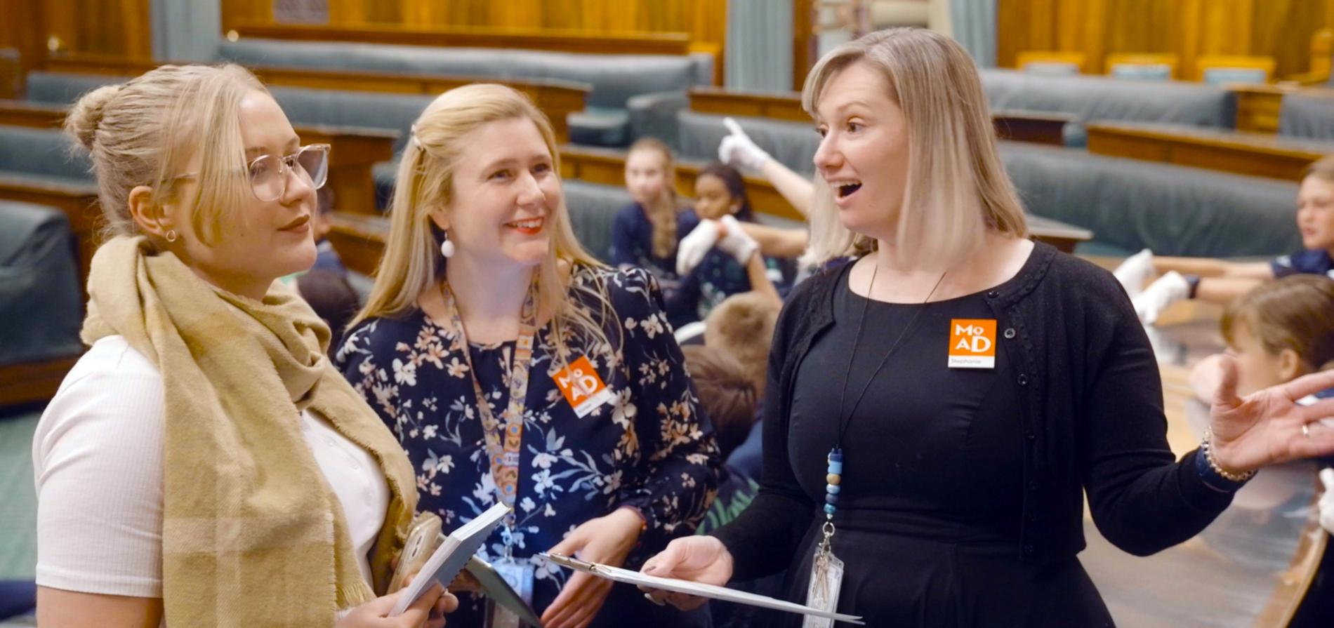 Two people stand holding clipboards, smiling and talking to a person about old parliament house's chamber of representatives.