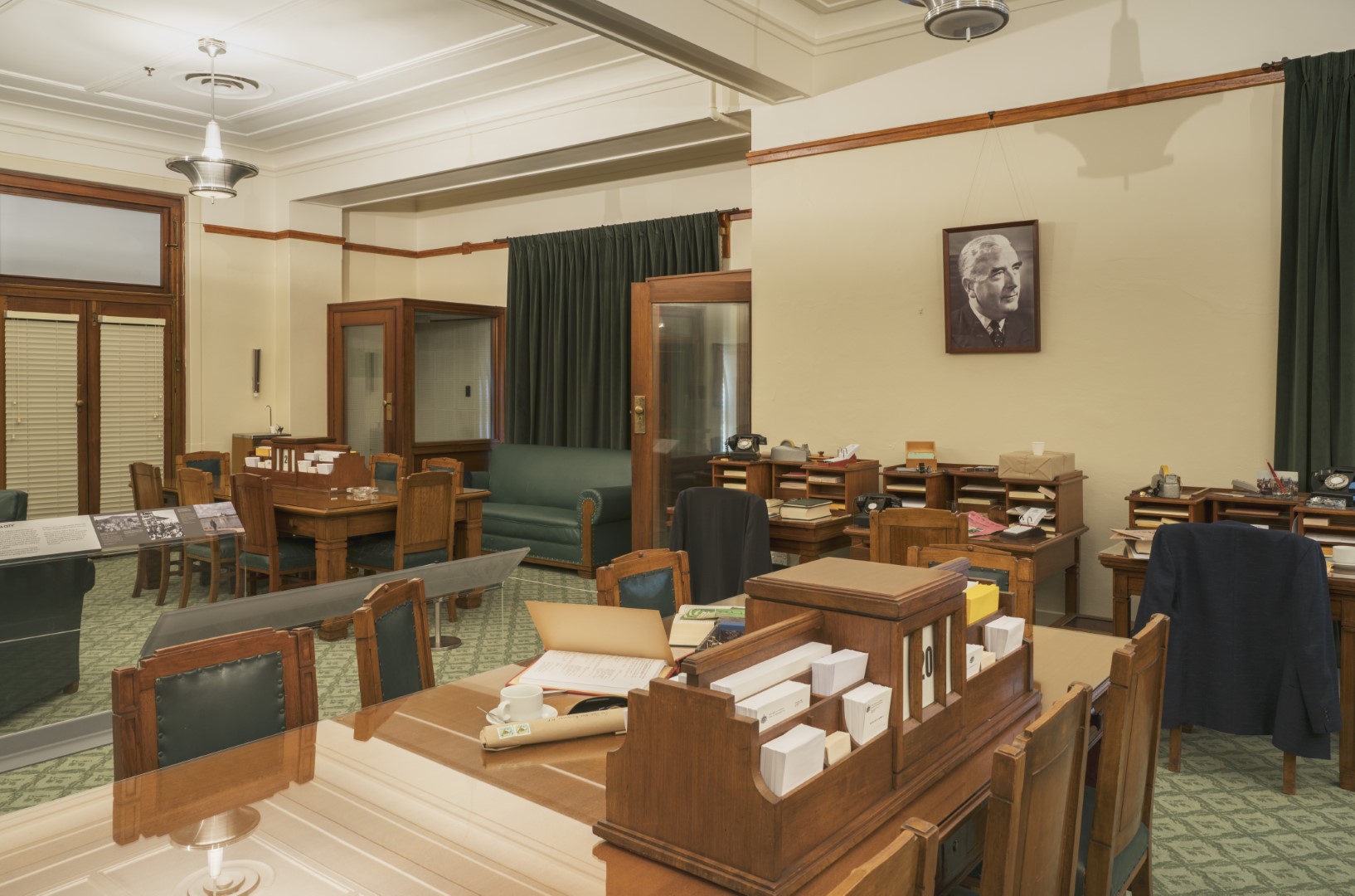 This colour photograph shows the Government Party Room re-created to evoke the atmosphere of 1964. There are two large meeting tables surrounded by chairs, three small desks, and two telephone booths. A large portrait of Prime Minister Robert Menzies is on one wall. The surfaces of the meeting tables and desks are covered with stationery, papers, telephones and ashtrays. This colour photograph shows the Government Party Room re-created to evoke the atmosphere of 1964. There are two large meeting tables surrounded by chairs, three small desks, and two telephone booths. A large portrait of Prime Minister Robert Menzies is on one wall. The surfaces of the meeting tables and desks are covered with stationery, papers, telephones and ashtrays.