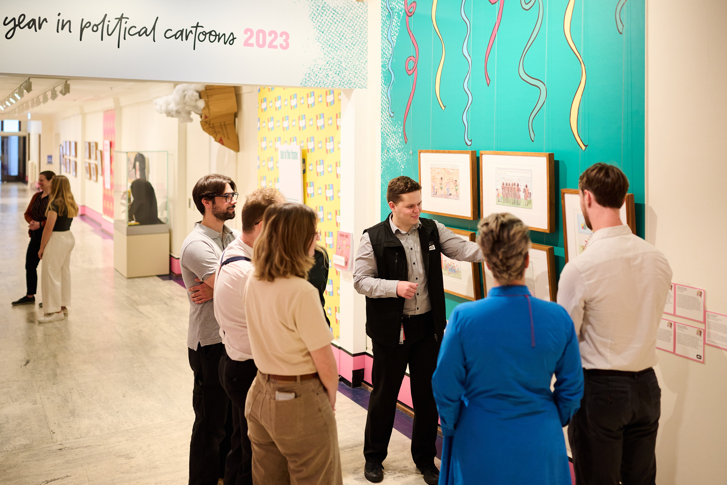 A tour guide wearing a MoAD badge shows a group of five people framed cartoons hanging in an exhibition.