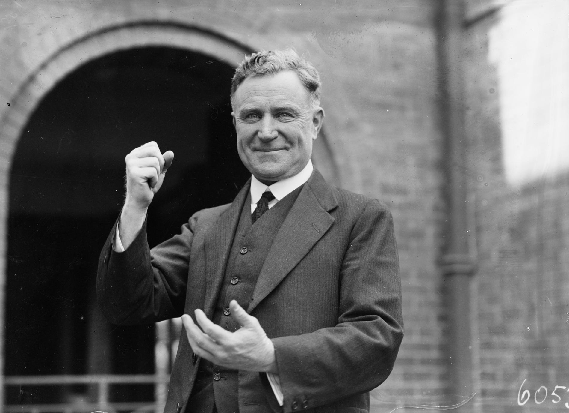 A man, Earle Page, wearing a striped three-piece suit, smiles and poses for a camera with a raised fist.