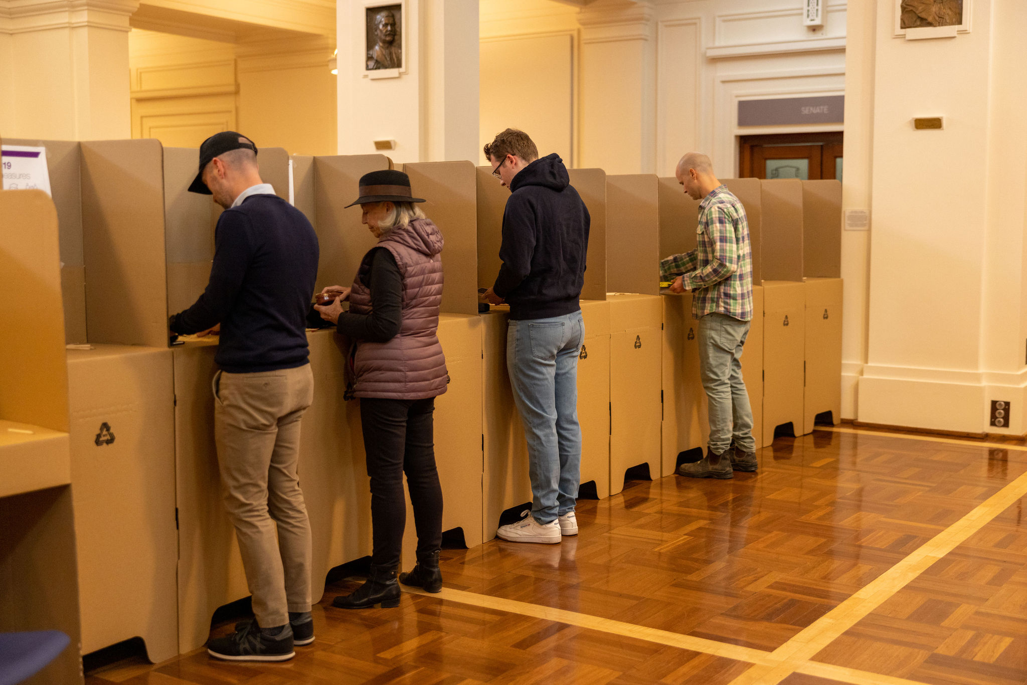 Four people, three men and a woman, standing at cardboard election booths in King's Hall.