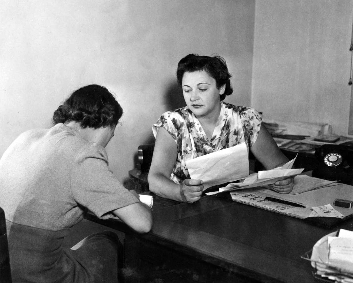 Nancy Wake is at a desk holding papers. She is looking across at another woman who is writing.