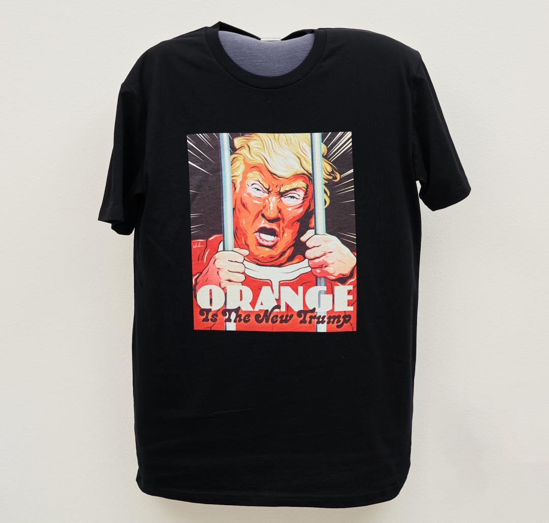 A black T-shirt with an image of an orange-faced Donald Trump wearing orange and looking angry, his hands cling to metal bars in front of him, text says, 'Orange Is The New Trump'.