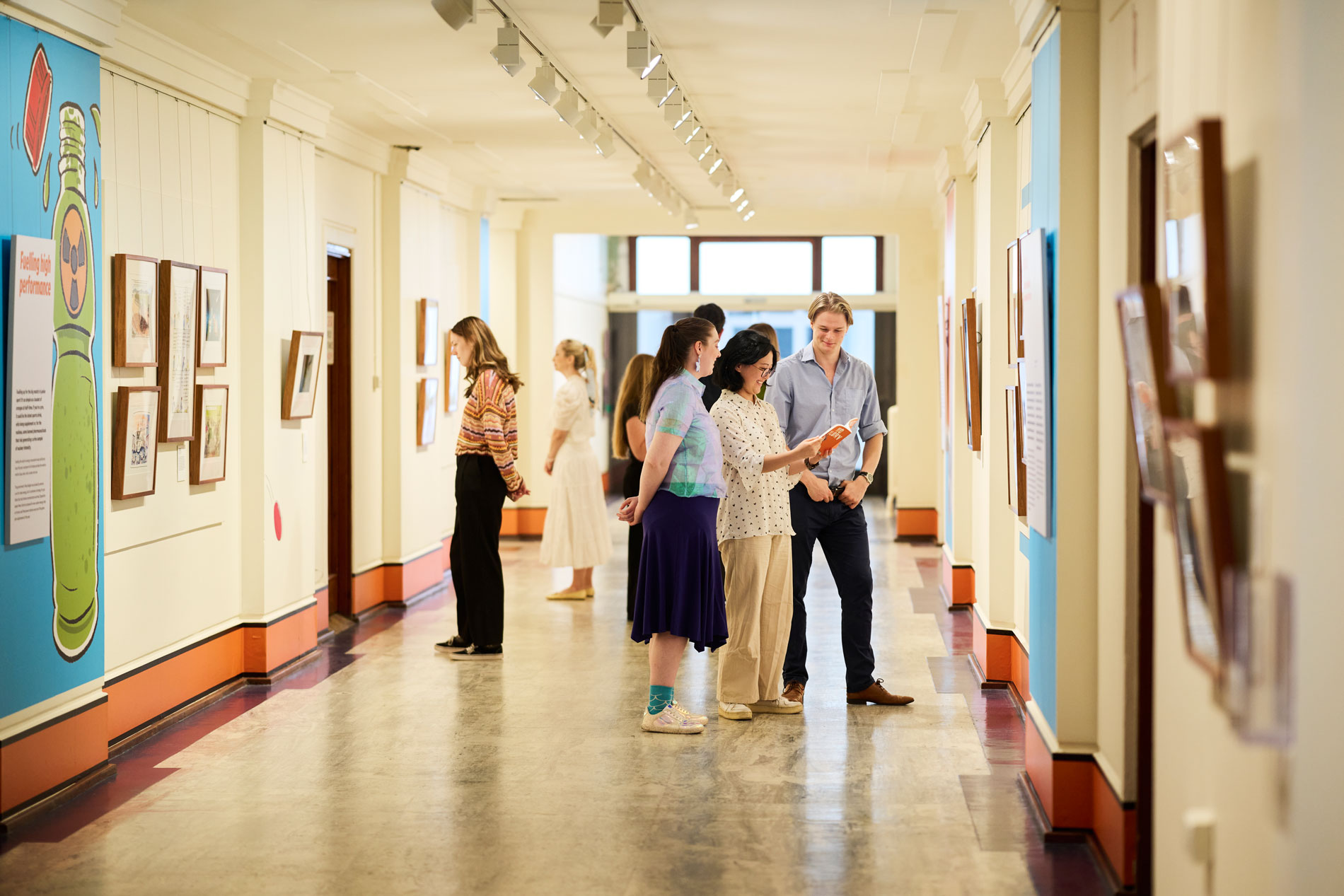 A crowd of people in a corridor of Old Parliament House look at a colourful exhibition of political cartoons.