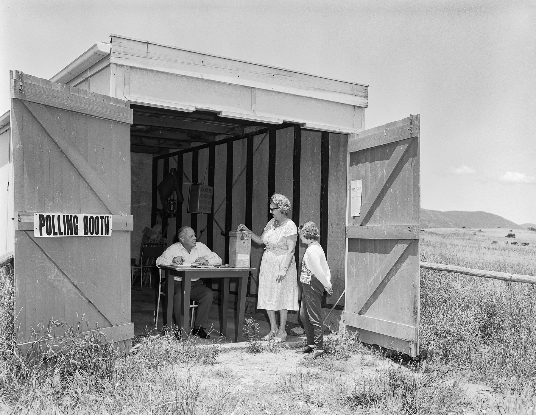 A woman puts a voting card in a ballot box at a shed in a paddock being used as a polling booth. A man sits at a table and a child looks on. 