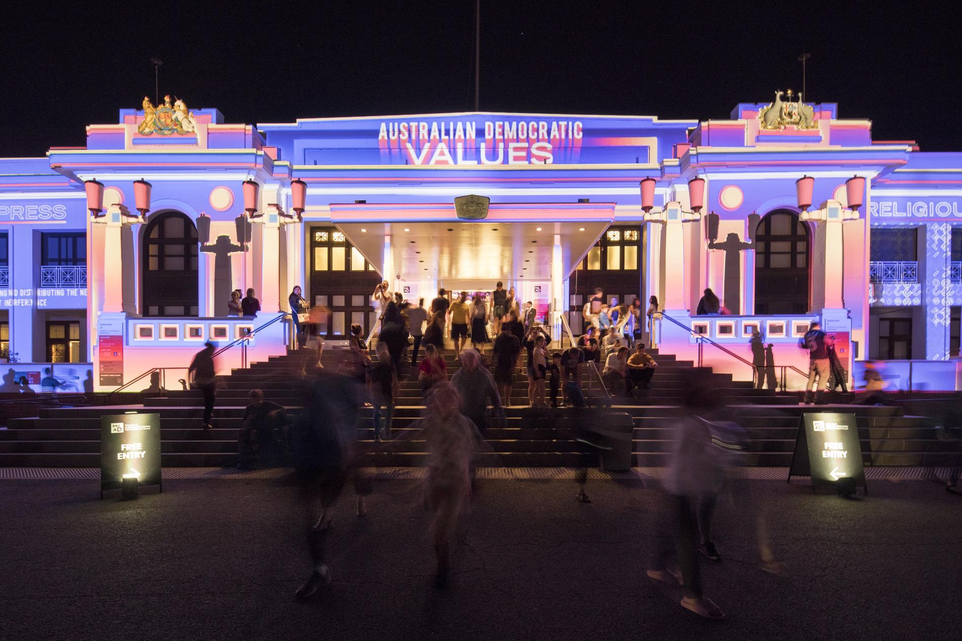 The front of Old Parliament House at night lit with multi-coloured lights and the words 'Australian democratic values'. A crowd of people walk up the stairs into the building.