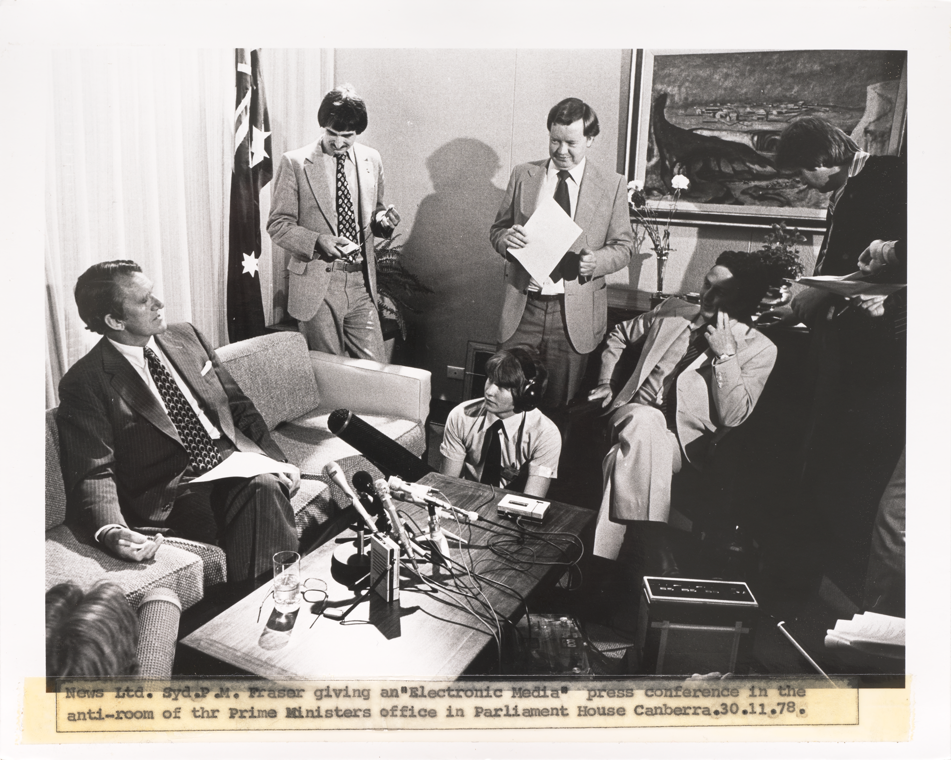 Prime Minister Malcolm Fraser sitting on a lounge in a room in Parliament House, with about 7 microphones pointed at him, surrounded by electronic media journalists.