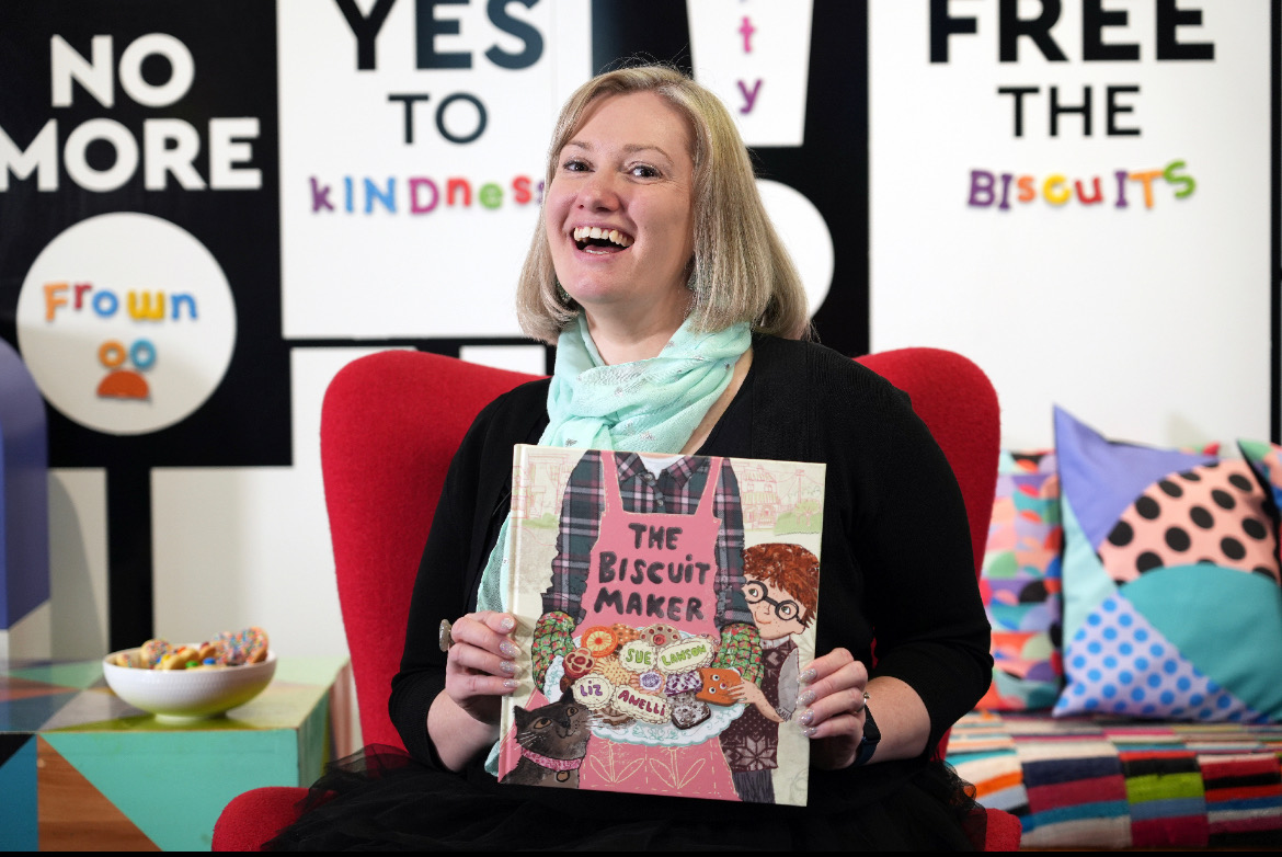 A woman sitting holding a picture book titled 'The Biscuit Maker' by Sue Lawson, illustrated by Liz Anelli