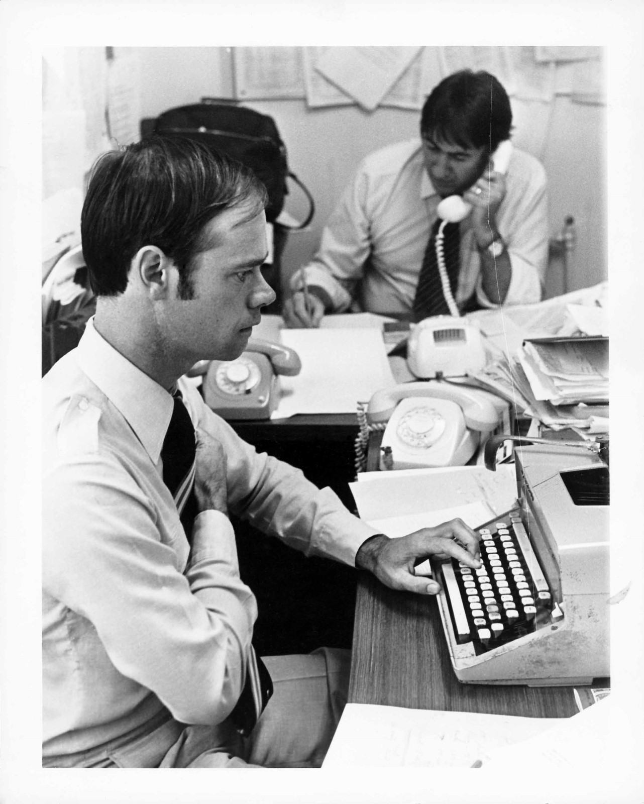 A man sitting at a desk, working on a typewriter in the Press Gallery while another man is on the phone behind him.