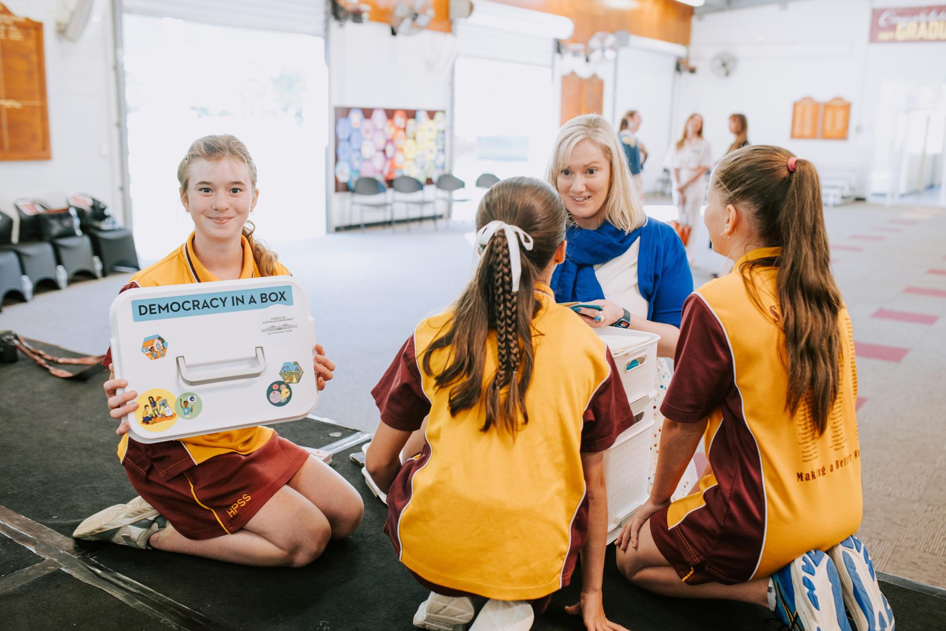 3 students in uniform engaging with Democracy in a Box while talking to a teacher.
