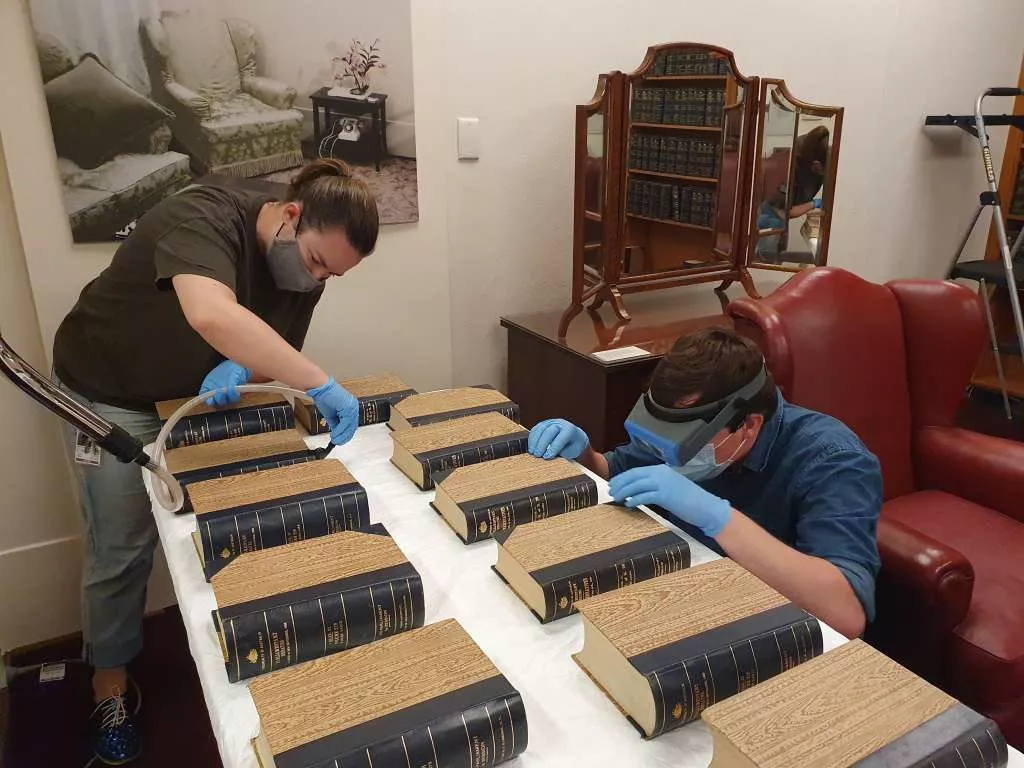 Two people lean over a table of old Hansard books and delicately clean them wearing gloves. 