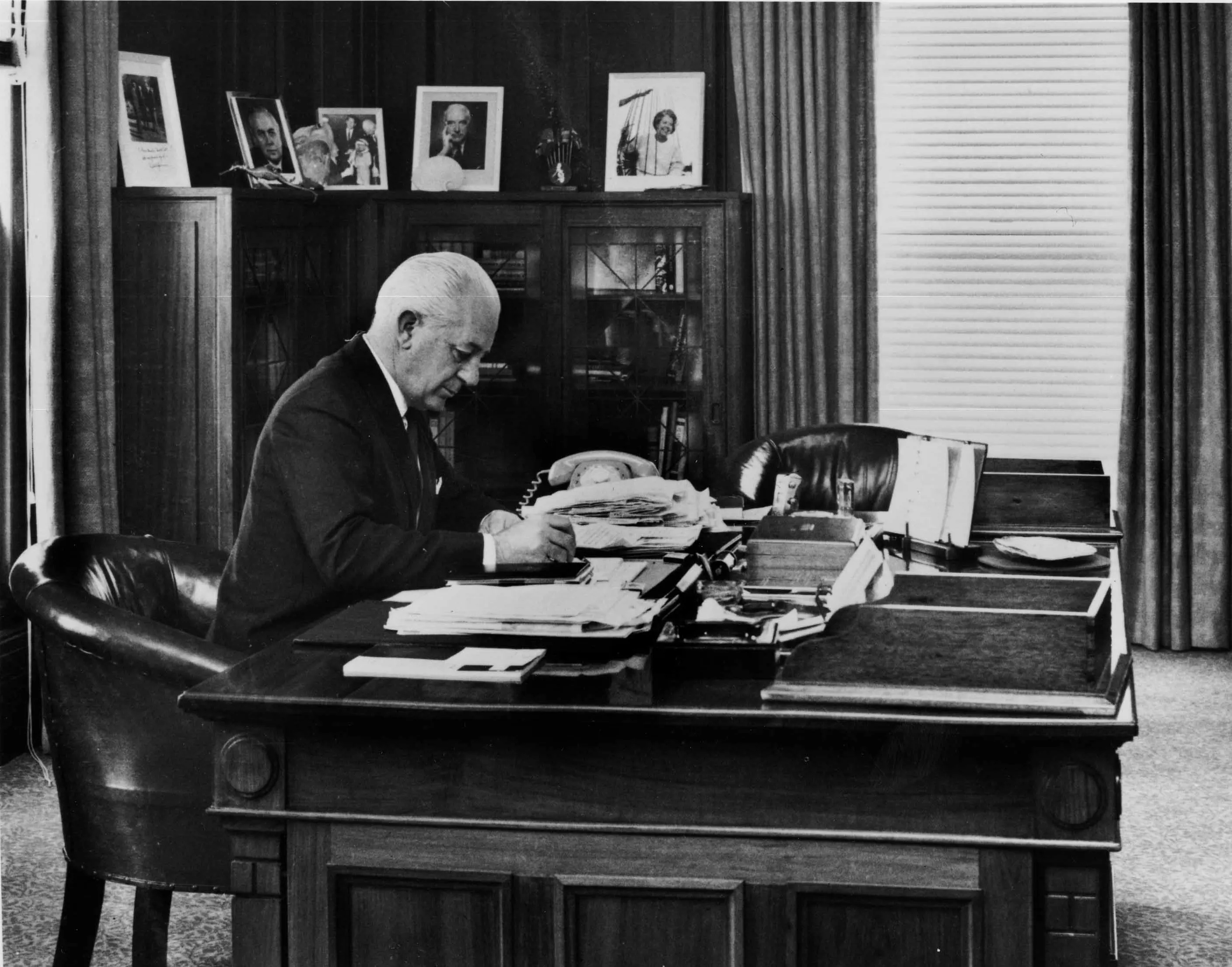 In this black and white photograph Prime Minister Harrold Holt sits at the timber desk made in 1926. Holt is wearing a dark suit, white shirt and tie and is leaning on the desk while he writes. The desk has fine carved panels with a geometric detailing on the corners. The surface of the desk is cluttered with stacks of documents and a telephone. On the glass-fronted cabinets in the background are a number of framed photographic portraits.  