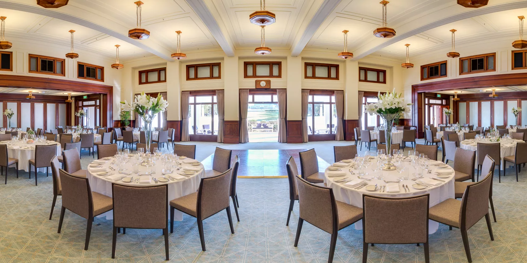 An art deco styled room with wooden pendant chandeliers, white ceilings and carpeted floor, with circular tables set with white tablecloths and lilies.