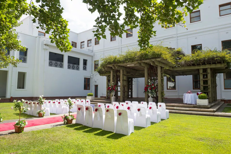White chairs set up in rows on a lawn facing a wooden pergola covered in vines, ready for a wedding.