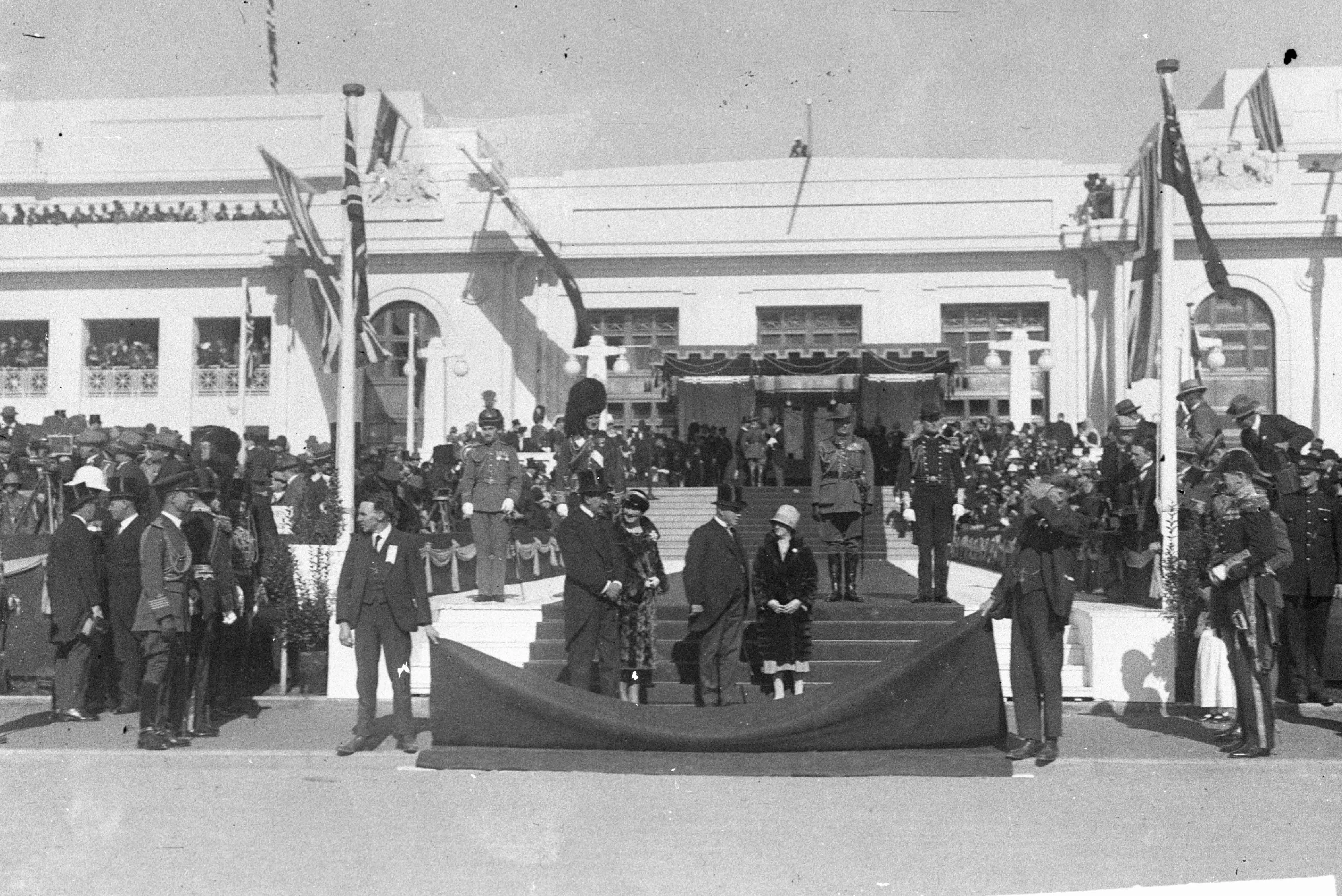 A black and white photo of the facade of Parliament House decorated with flags, a canopy and welcome carpet before the arrival of the Duke and Duchess of York for the opening of the building. There are crowds on the roof, front steps, verandahs and flanking the front door. A guard of honour in military uniform stands ready. Two attendants are holding up the end of the red carpet.