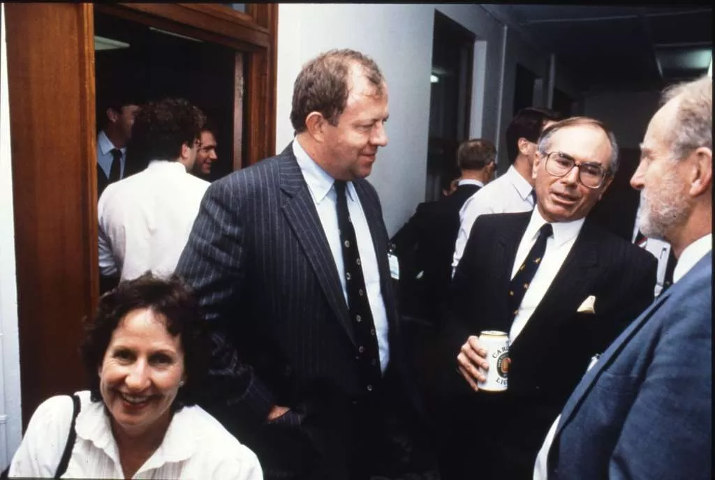 Opposition Leader John Howard, wearing a suit and tie, has a beer with colleagues in a crowded room.