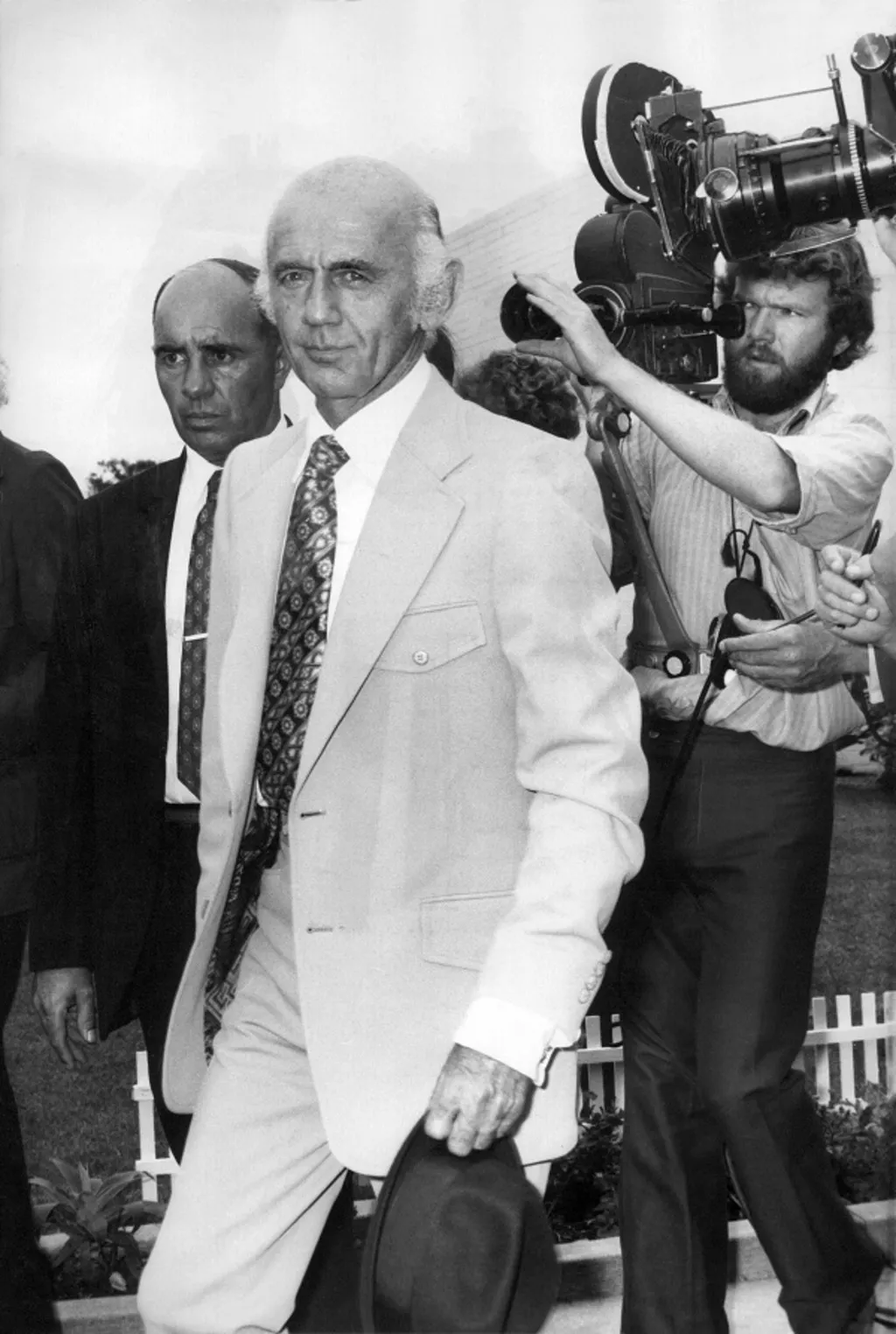 Billy McMahon wearing a light-coloured suit, hat in hand, followed by a film camera operator.