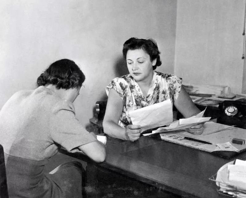 Nancy Wake, a short-haired woman, sitting at a desk, holding papers in her hand.