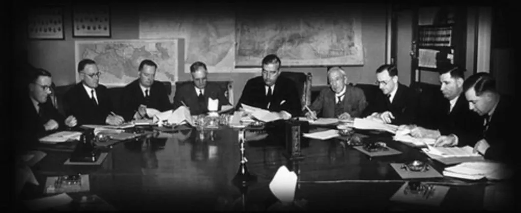 Prime Minister Robert Menzies sits at a table, with four men sitting on each side, all looking at papers, large maps pinned to the wall in the background.