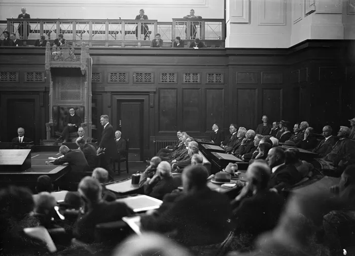 A black and white photo of the House of Representatives Chamber with men sitting in rows on leather chairs in a U-shaped pattern. A large ornate chair at the front of the room has a man sitting in it. 