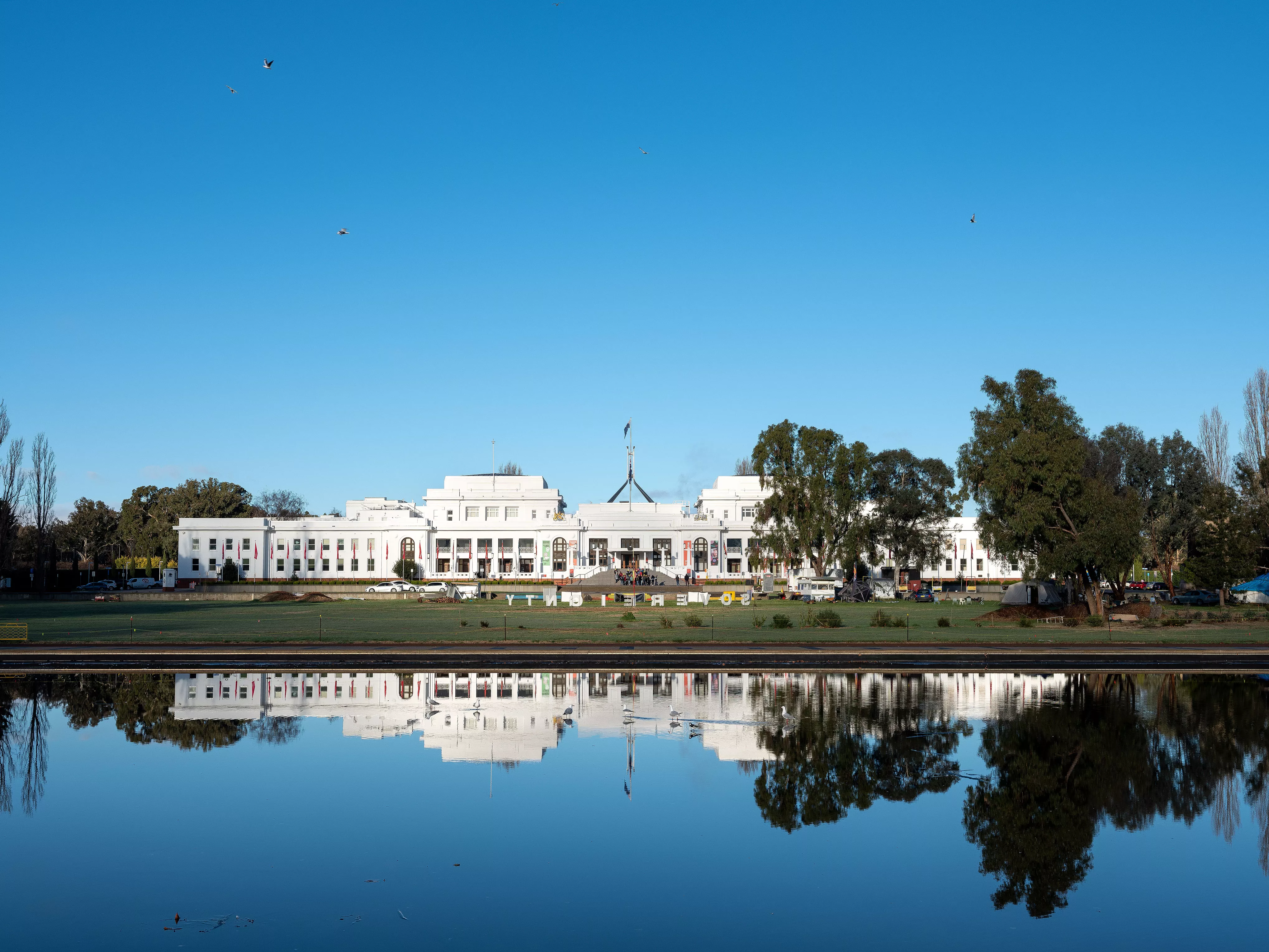 A view from across a lake looking over to Old Parliament House, a long white building with a flag on top of it. The Aboriginal Tent Embassy is in front of this. 