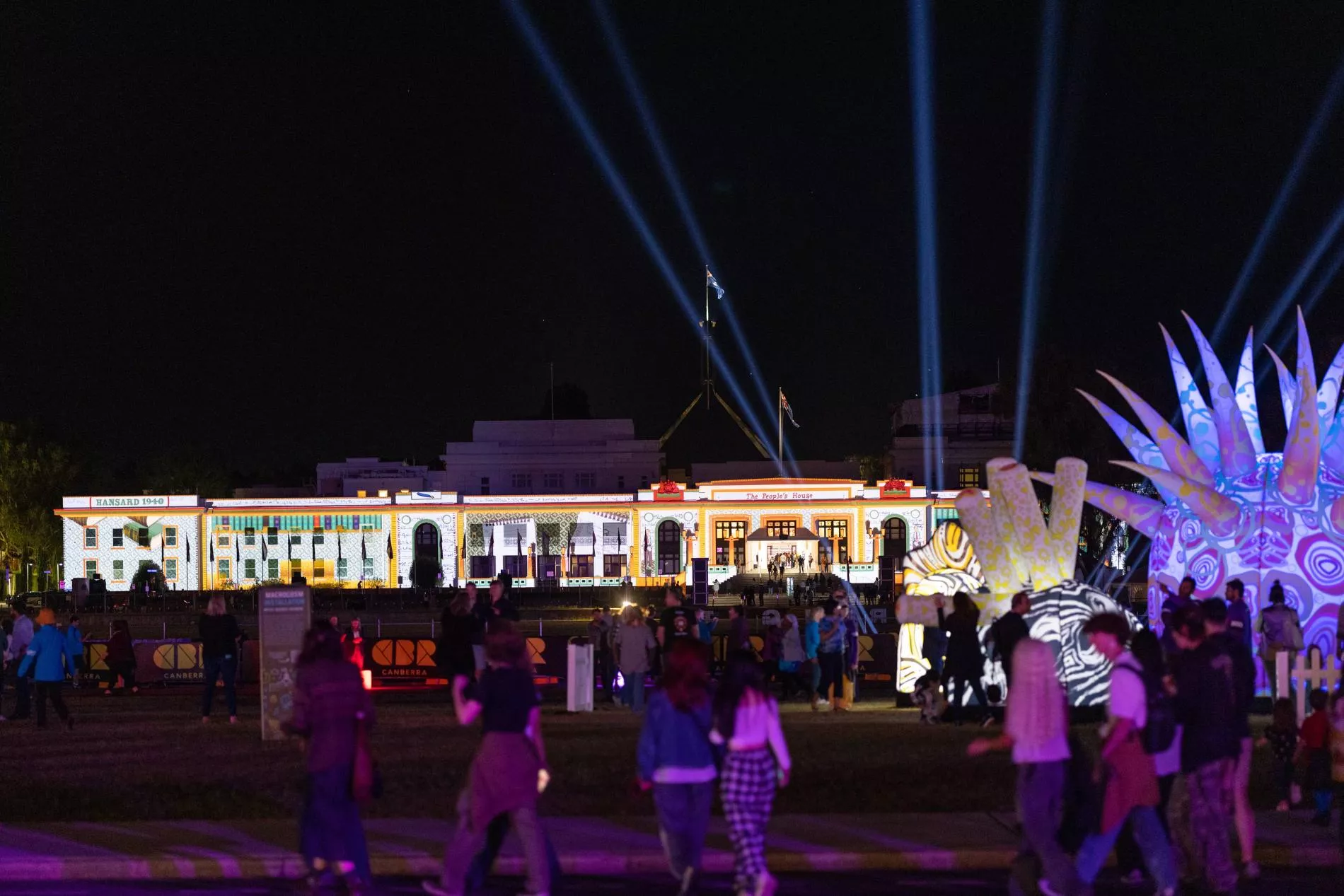 A night shot of Old Parliament House, a large white long building, with people gathering out the front and coloured lights projected onto the surface.