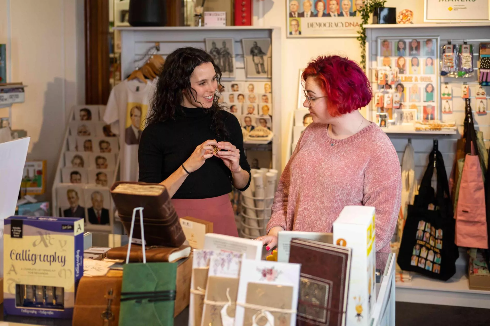 Two women talk to each other in a shop surrounded by books, postcards and bags featuring prime ministers. 