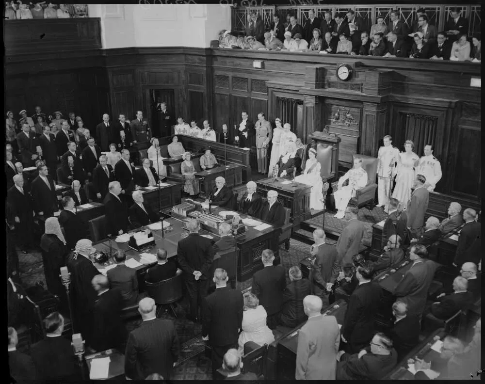 A black and white photo of the Senate Chamber filled with people standing up looking to an ornate chair at the front of the room where the Queen sits wearing a crown and a sash.