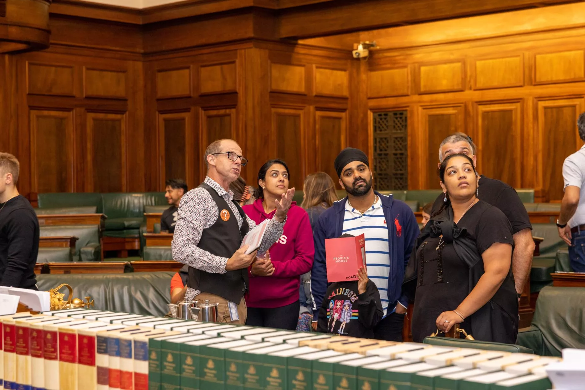 A tour guide points to something above a group of people listening to his tour. They are in the House of Representatives Chamber with green leather chairs behind them and leather bound books stacked on a central table in front of them.