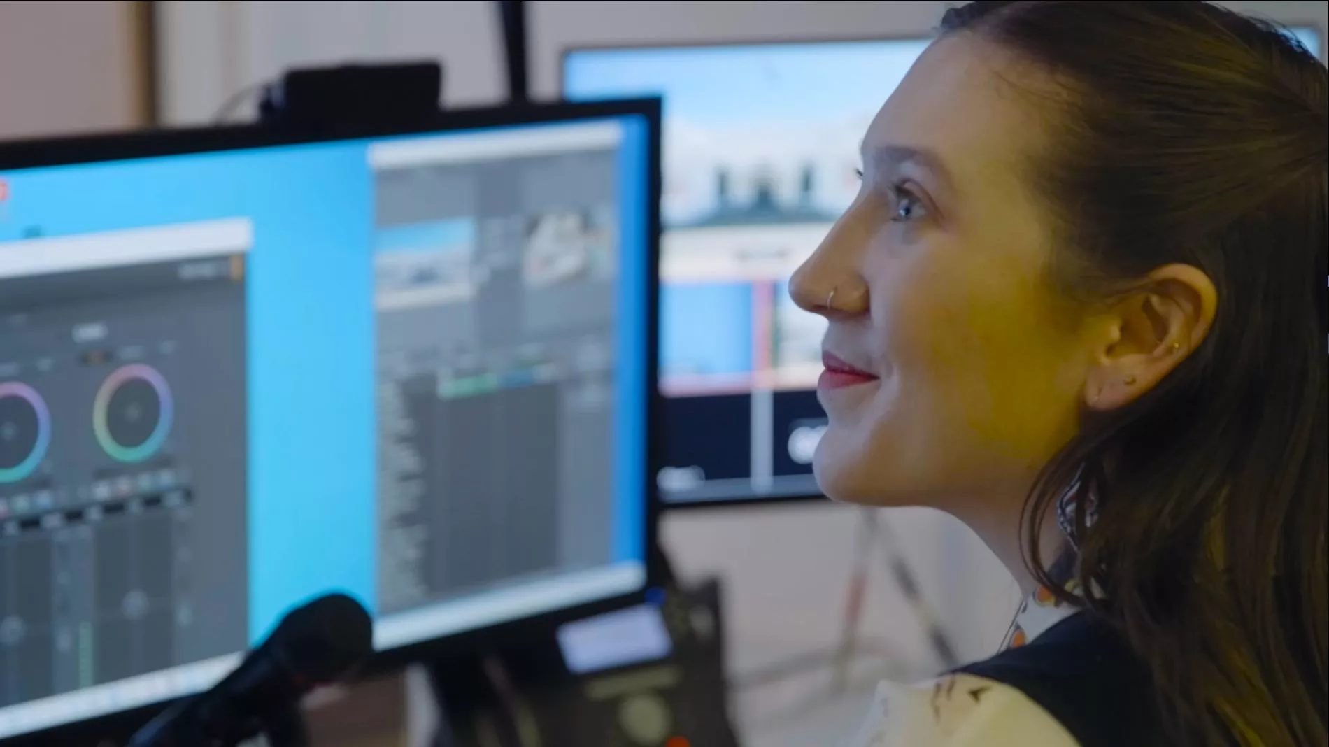 A woman looks off to the side and some screens and other technology are out of focus in the background