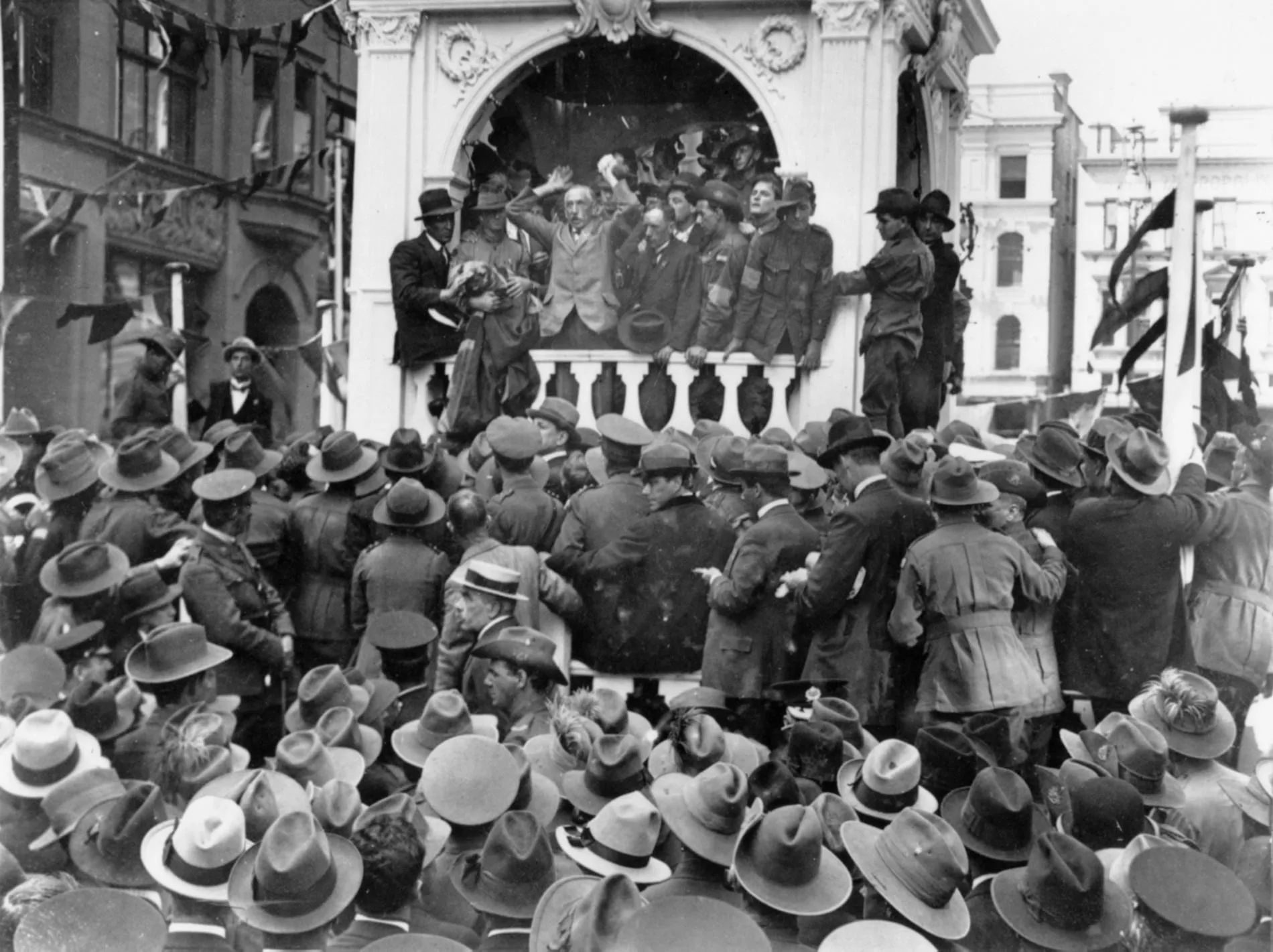 A black and white photo of large group of people wearing hats crowd around a podium where Billy Hughes is giving a speech. 