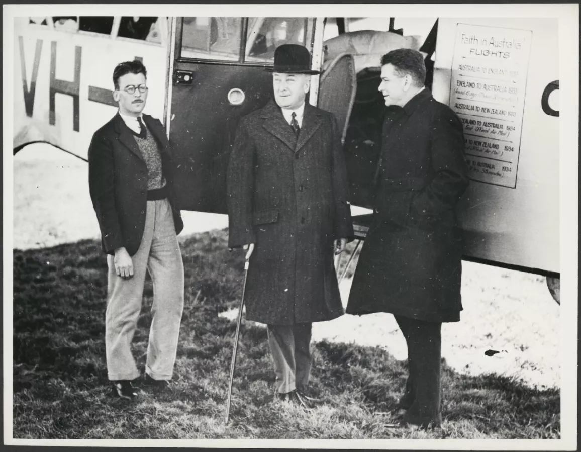 Aviator Charles Ulm and Prime Minister Joe Lyons stand with another man in front of an airplane. It's a black and white photo.