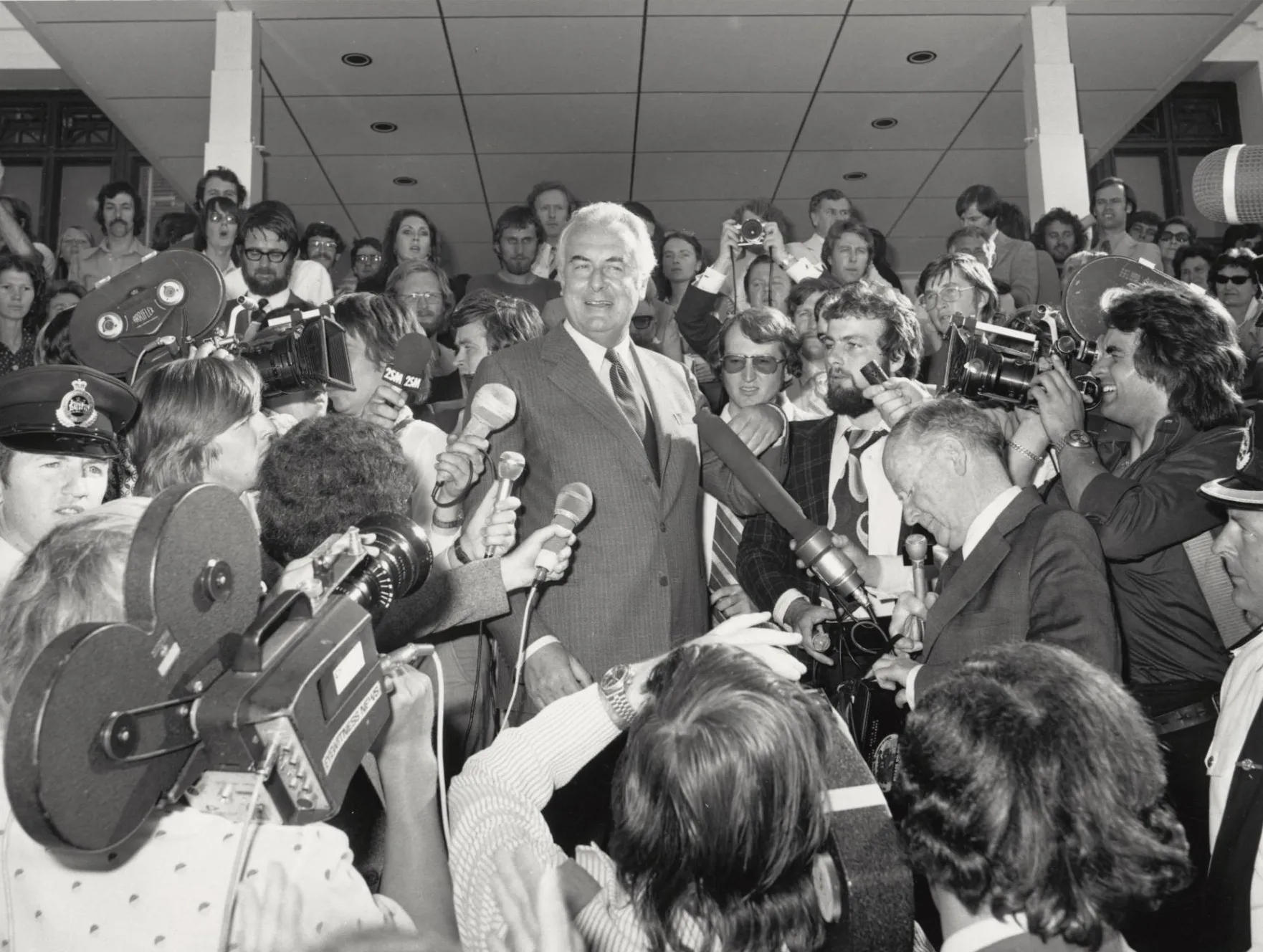 Gough Whitlam on the steps of Old Parliament House surrounded by the press who are holding microphones and video cameras on 11 November 1975.