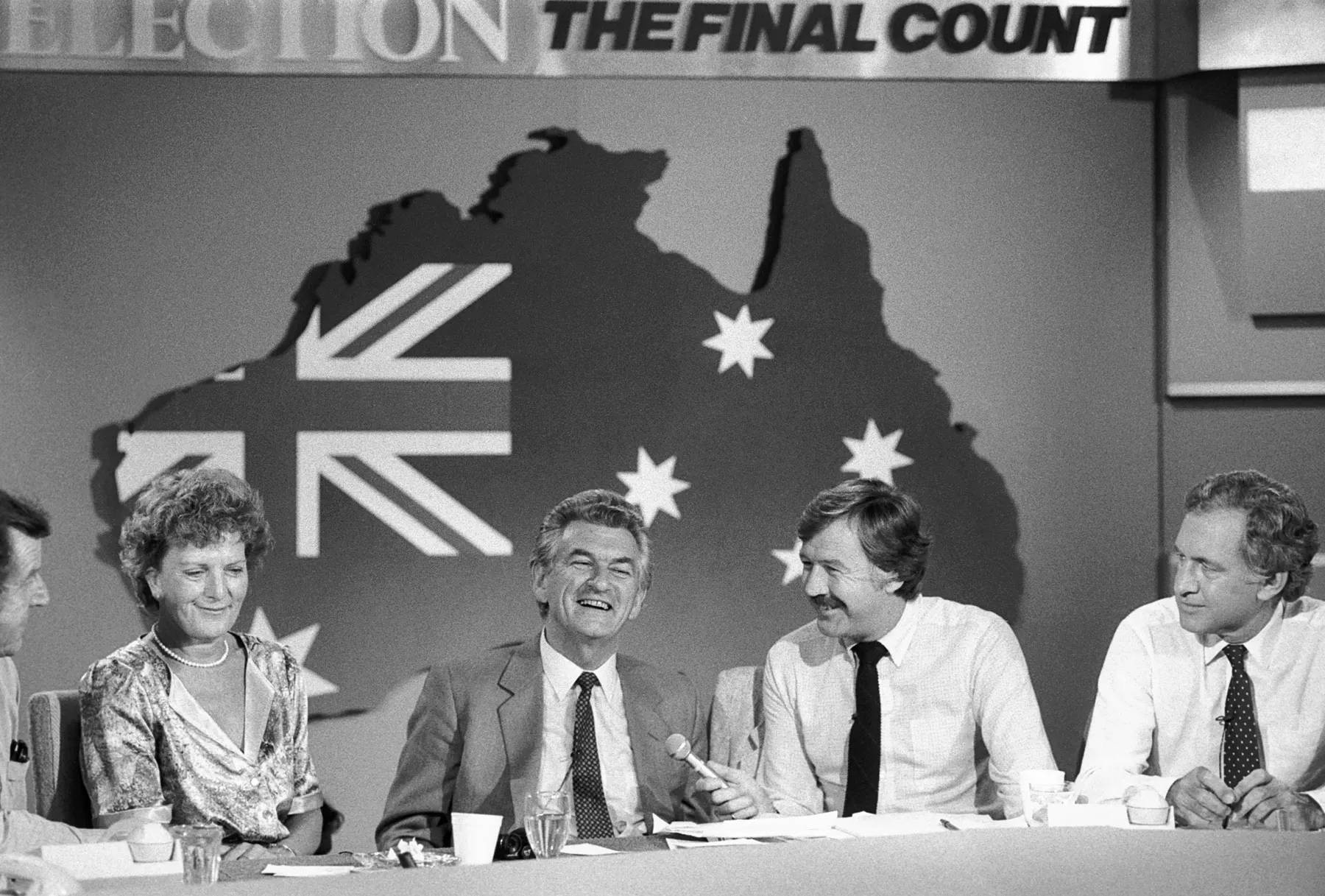 Hazel and Bob Hawke sit with three journalists at a table in the National Tally Room in 1983. There is a large map of Australia behind them on the wall.