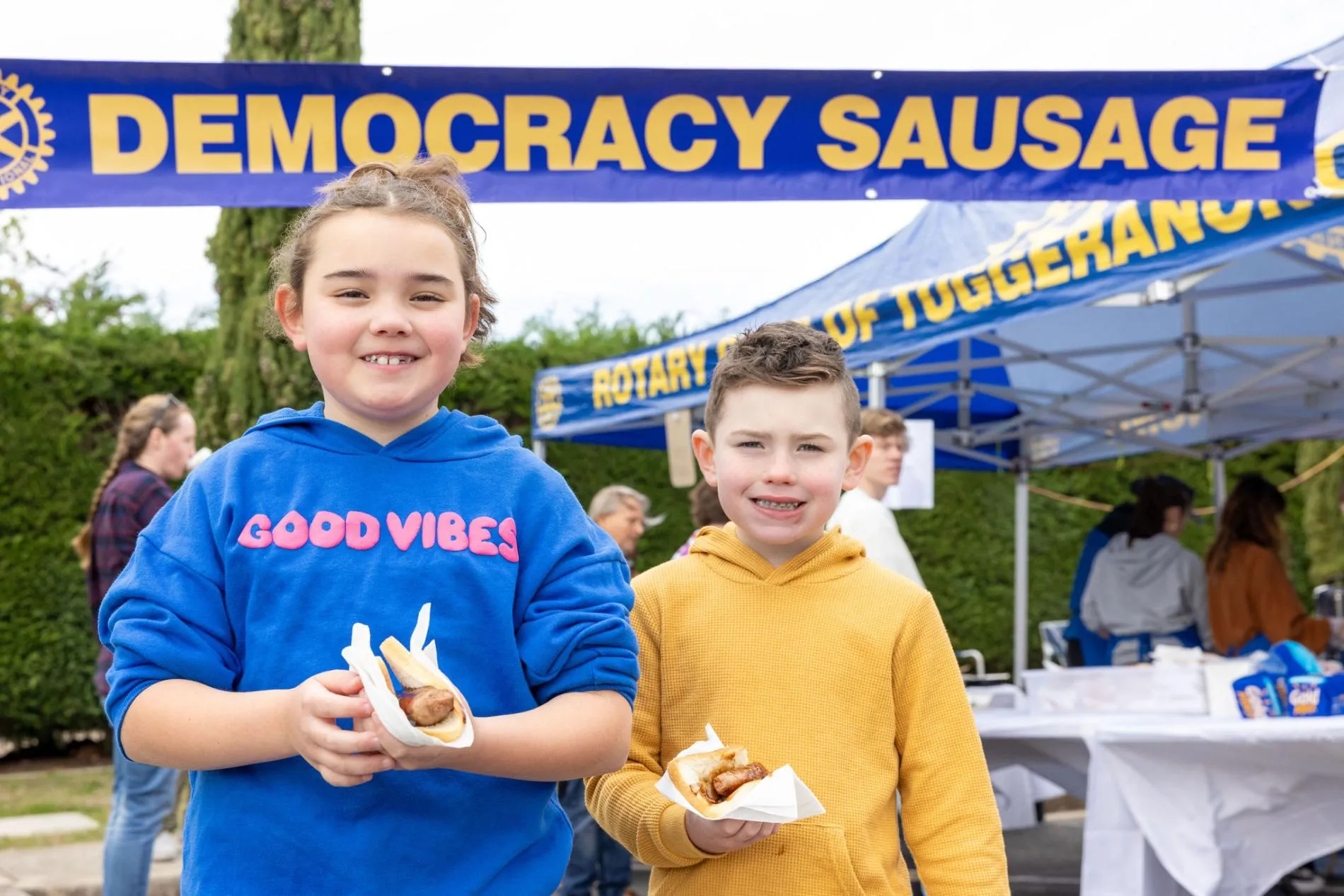 A girl wearing a blue hoodie with the text 'Good vibes' and a boy wearing a mustard hoodie each hold a sausage sandwich in front of an outdoor tent with a 'Democracy sausage' banner in the garden of Old Parliament House.