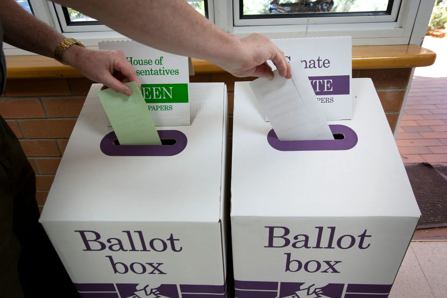 Hands place green and white ballot papers into white and purple ballot boxes.