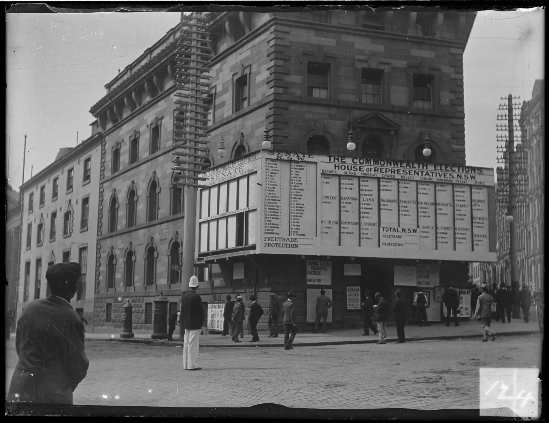 People stand on the street in Sydney looking at a building displaying a giant tally board showing the results of the 1901 federal election.