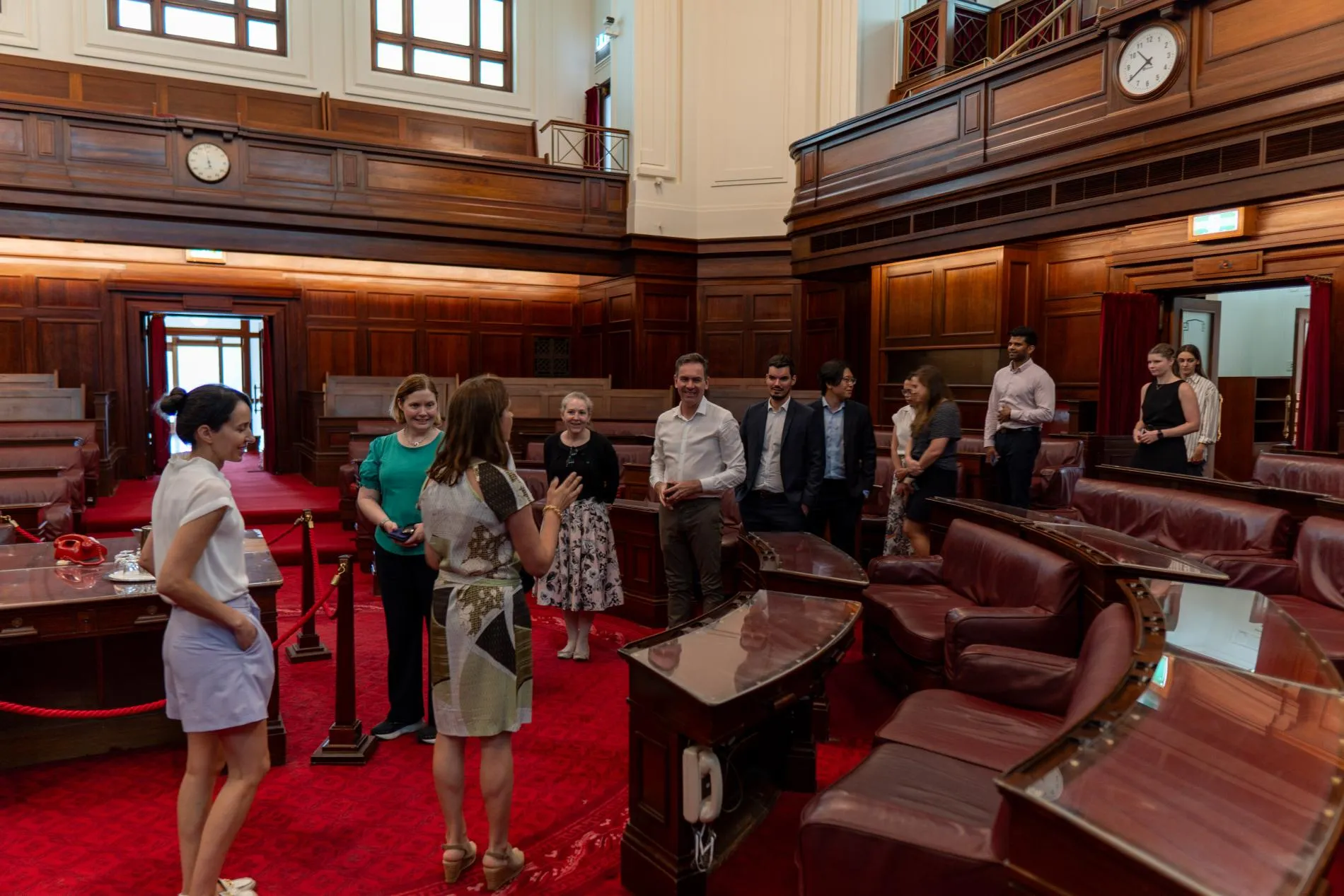 People on a tour in the Senate Chamber at Old Parliament House, it has red carpet and leather bench seats.